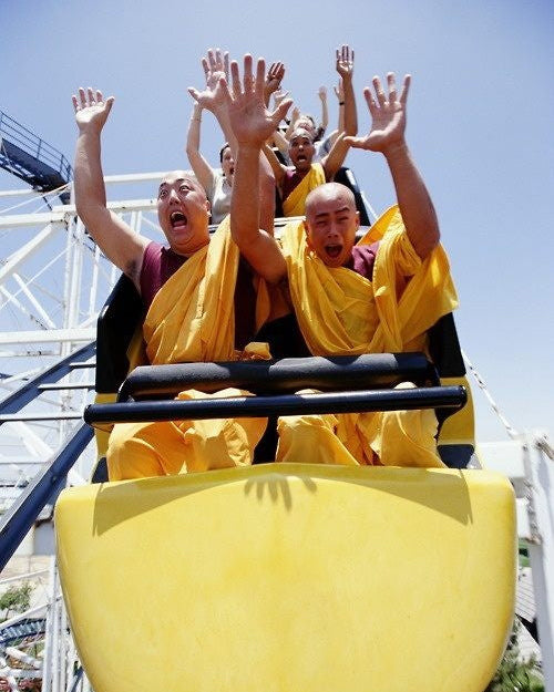 Monks in yellow robes on a roller coaster with arms raised against a blue sky.